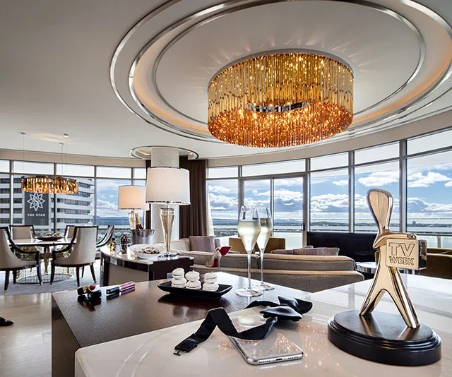 Luxurious hotel room with elegant chandelier, TV Week Logie award, wine glasses, and panoramic views of the city.
