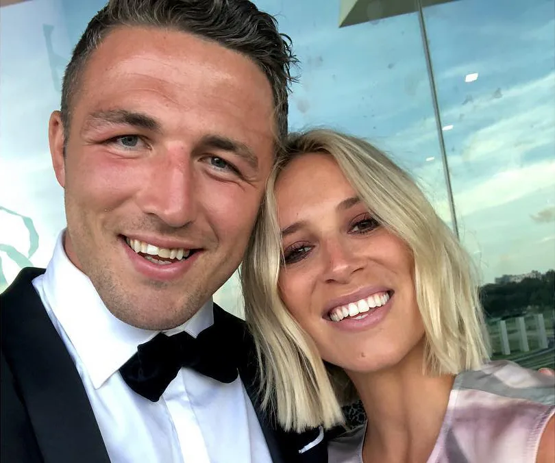 Two people smiling for a selfie, dressed formally with a blurred outdoor background.