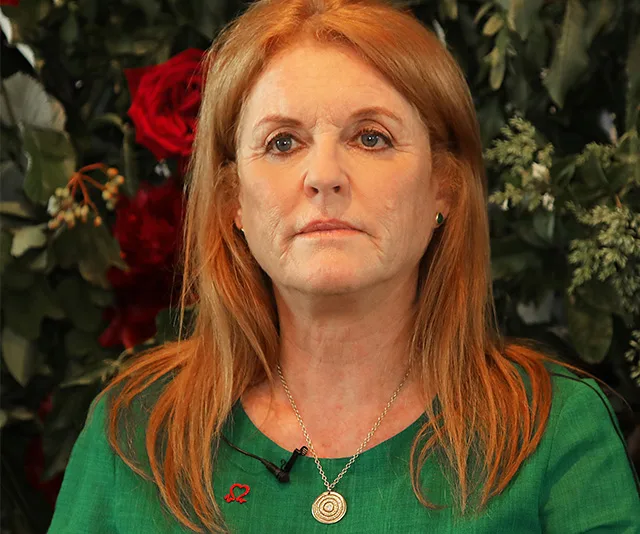 Sarah Ferguson in green, wearing a necklace, speaks in front of a floral backdrop at a charity event.