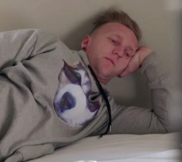 A person in a gray hoodie with a dog's face, lying on a bed, leaning on one elbow, looking contemplative.