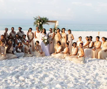 Bride and 27 bridesmaids in light beige dresses pose on a sunny beach with a wooden arch in the background.