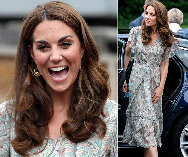 A woman in a patterned dress smiles broadly, standing beside a car, her hair styled in loose waves.