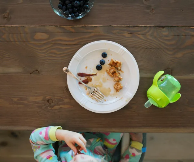 Child's breakfast with waffles, blueberries, bacon, and a green sippy cup on a wooden table.