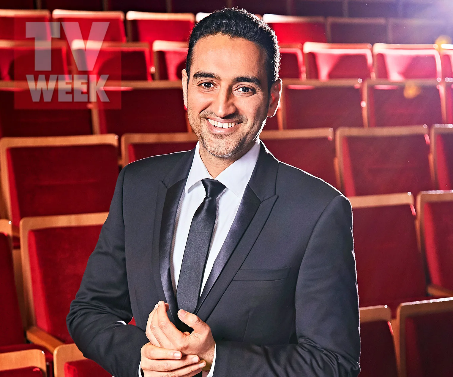 A person in a suit smiling in an empty theater with red seats, under TV WEEK logo.