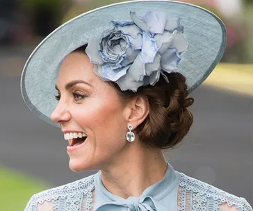 A woman smiling, wearing a light blue dress and matching hat with a large flower decoration.