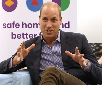 A man in a suit gestures while speaking, seated in front of a sign reading "safe homes and better f..."