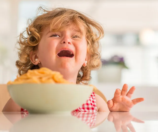 A child in a red checkered dress crying at a table with a bowl of pasta in front of her.