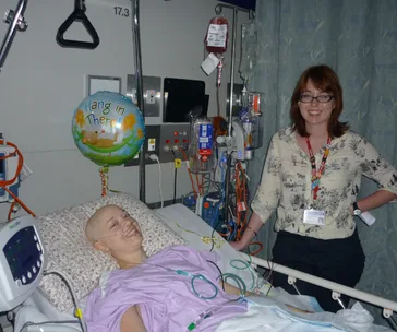 A patient in a hospital bed with IVs and monitors nearby, accompanied by a woman standing beside. Balloon reads "Hang in There."