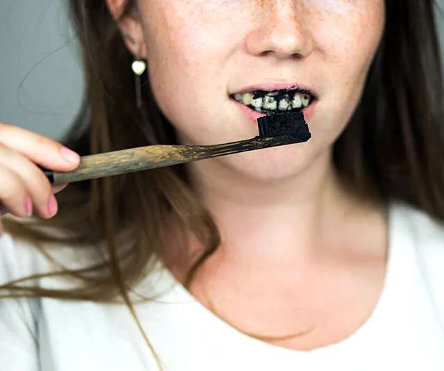 A person brushing teeth with charcoal-based toothpaste, showing blackened mouth.