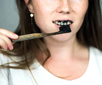 A person brushing teeth with charcoal-based toothpaste, showing blackened mouth.