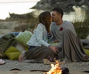 A couple kissing near a campfire by a lake, wrapped in blankets on a picnic setup.