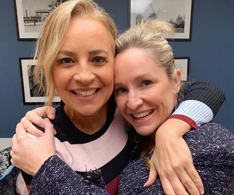 Two women smiling warmly, embracing each other in a cozy setting with framed pictures on the wall behind them.