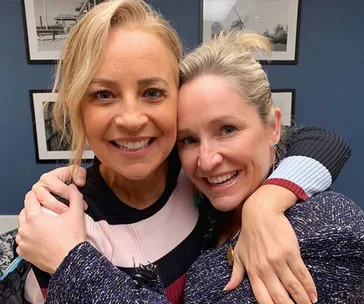 Two women smiling warmly, embracing each other in a cozy setting with framed pictures on the wall behind them.