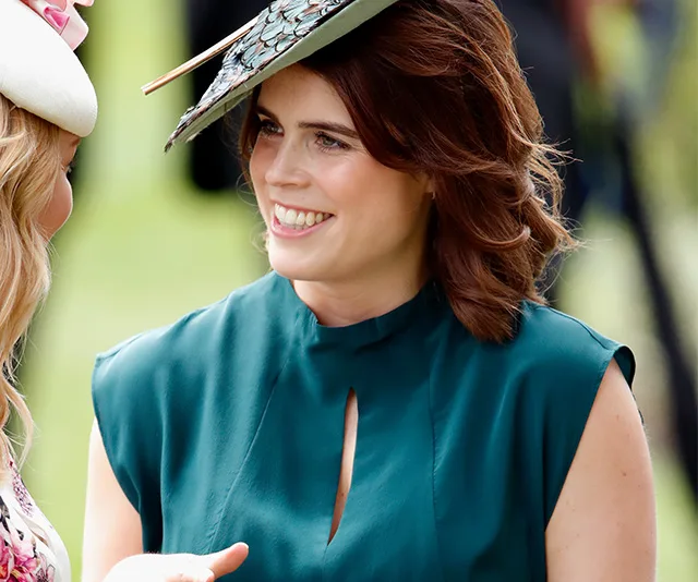 A woman in a teal dress and decorative hat smiles at an outdoor event.