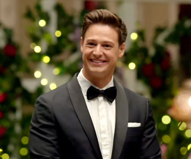 A man in a tuxedo smiling, set against a backdrop of blurred lights and greenery.