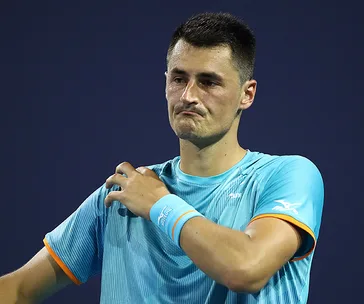 Tennis player in a blue shirt looking disappointed, adjusting his collar on a dark background.