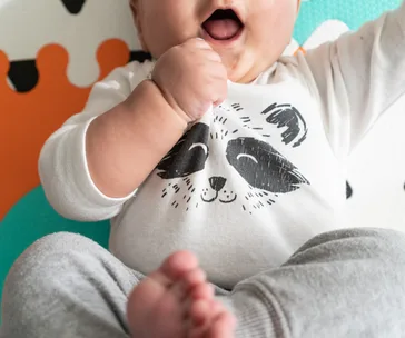 Baby wearing a white onesie with a raccoon print, sitting on a colorful activity mat with patterned design.