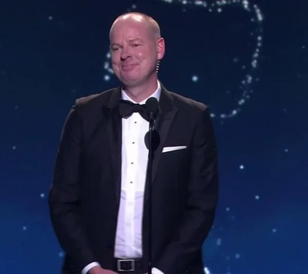 A man in a tuxedo speaks at a microphone on a stage with a starry background.