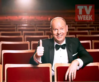 Comedian in tuxedo gives thumbs up in empty theater, "TV Week" logo visible in corner.