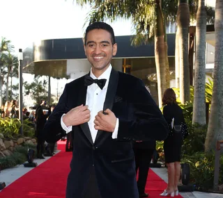 A man in a tuxedo smiles while standing on a red carpet with palm trees and a modern building in the background.