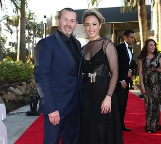 A man and woman pose together on a red carpet. They are dressed in formal evening attire.