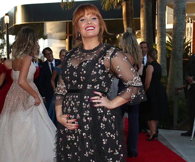 Person in a floral dress smiles and poses on a red carpet event, surrounded by attendees in formal attire.