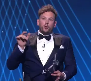Man in a suit holding an award, speaking on stage with a blue, patterned background.