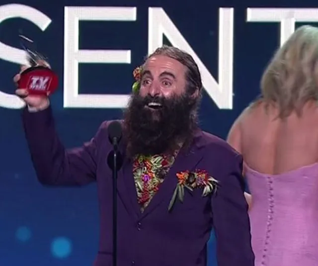 A man with a bushy beard in a floral shirt and dark suit joyfully holds an award on stage, smiling widely.