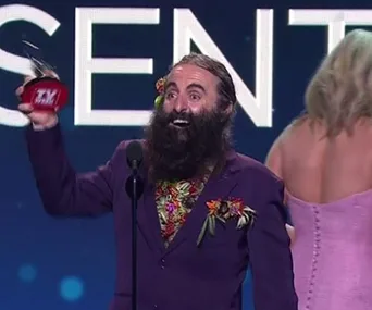 A man with a bushy beard in a floral shirt and dark suit joyfully holds an award on stage, smiling widely.