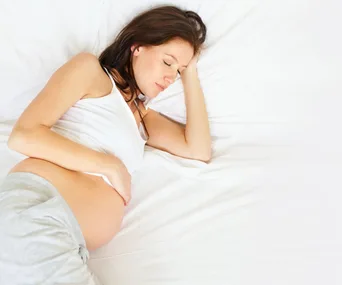 Pregnant woman sleeping on her side in a bed, wearing a white top and gray pants.