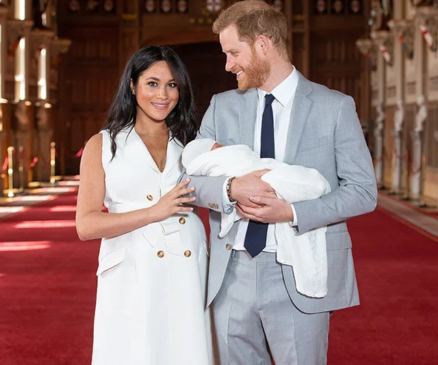A couple in formal attire holds a newborn wrapped in a white blanket inside a grand hall.