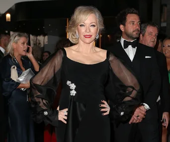 Elegant woman in a black dress poses confidently at an event with people in the background.