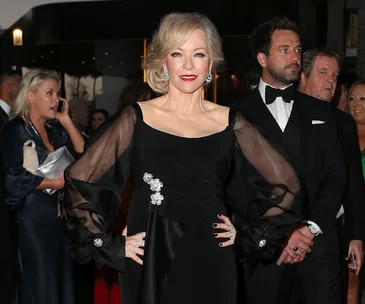 Elegant woman in a black dress poses confidently at an event with people in the background.