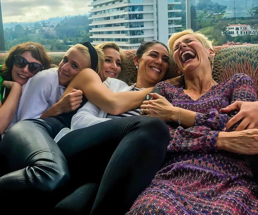 Group of five women sitting on a couch, laughing and embracing, with cityscape and buildings in the background.