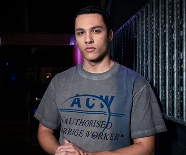 Young man with braided hair and a serious expression, wearing an "ACW" t-shirt, stands in an industrial setting.