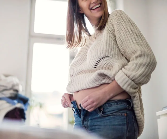 Pregnant woman smiling while adjusting jeans, showing a slight baby bump in a cozy home setting.