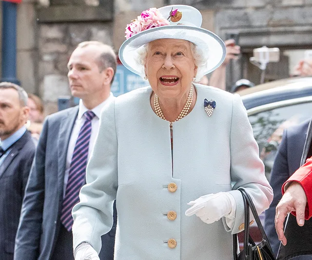 A joyful elderly woman in a light blue outfit and floral hat, smiling, surrounded by people outdoors.