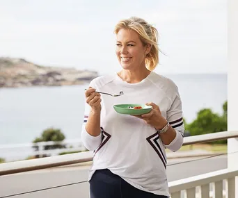 Woman in white sweater holding a bowl on a balcony, seaside view in background.