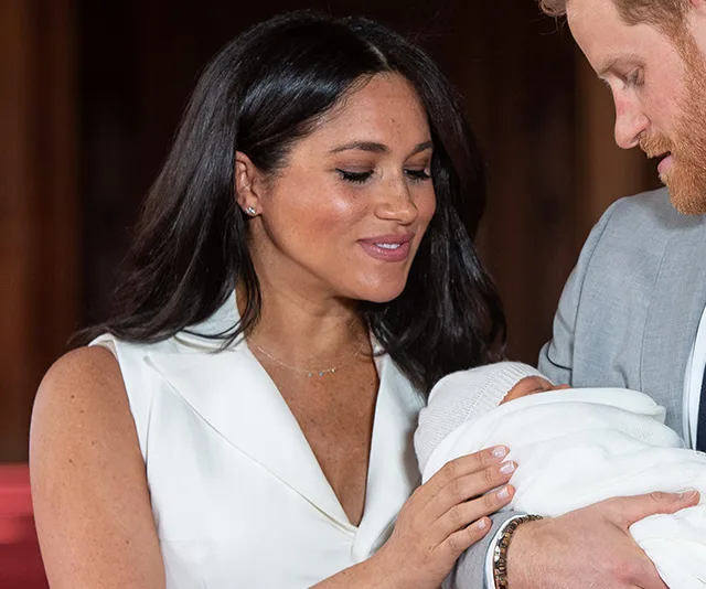 A woman in a white dress and a man holding a newborn wrapped in a white blanket, with a warm interior backdrop.