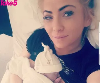 A woman lying down, smiling, holding a newborn baby wrapped in a white blanket wearing a hat.