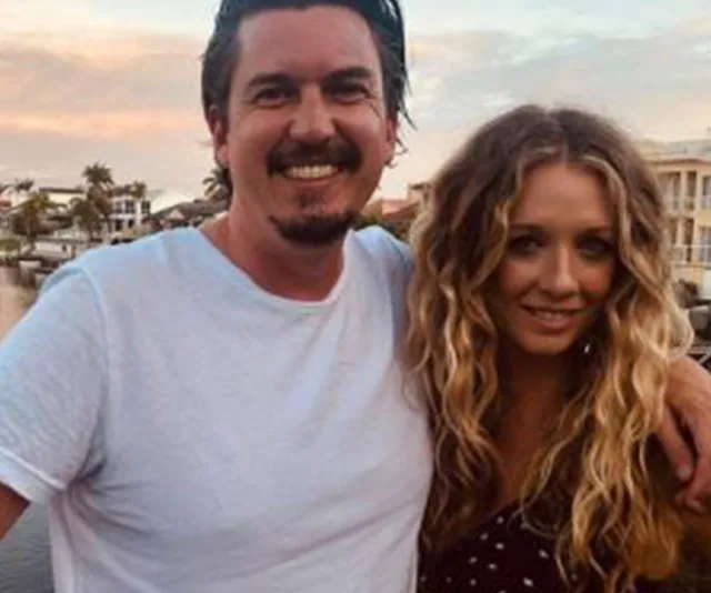 A smiling man in a white T-shirt with a woman with long wavy hair, standing by a waterfront at sunset.