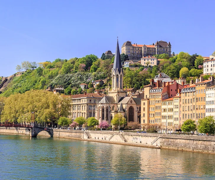 Riverfront view of Lyon, France with colorful buildings and a church spire under a clear blue sky.