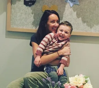 Woman holding a smiling child in a striped sweater, surrounded by flowers indoors.