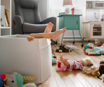 Child's legs sticking out of a toy bin surrounded by dolls and toys in a playroom.