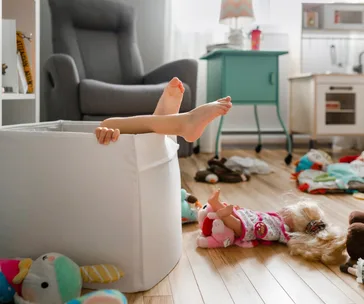 Child's legs sticking out of a toy bin surrounded by dolls and toys in a playroom.
