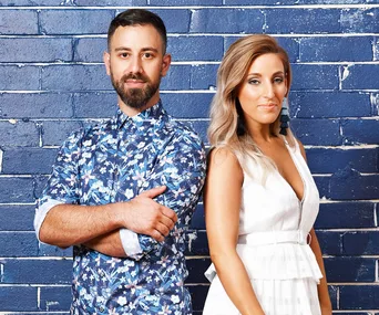 Two people standing against a blue brick wall, one in a floral shirt, the other in a white dress, both looking at the camera.
