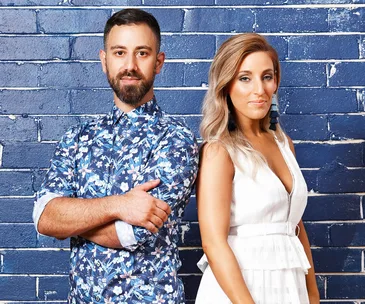 Two people standing against a blue brick wall, one in a floral shirt, the other in a white dress, both looking at the camera.