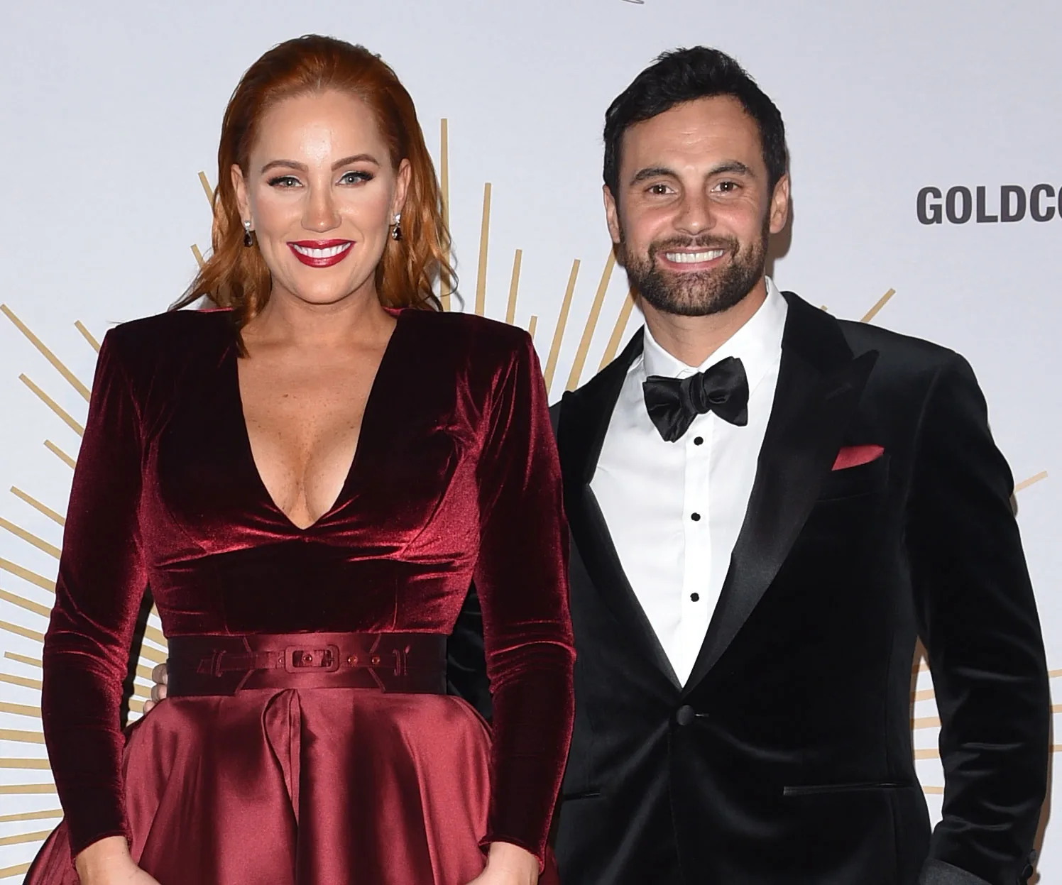 A couple dressed in formal attire, with the woman in a red velvet dress and the man in a black tuxedo, smiling at an event.