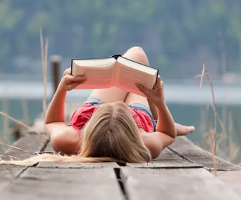 Young woman lying on a dock, reading a book with a lake and trees in the background.