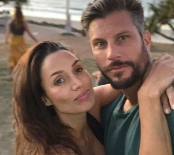 Couple posing outdoors near the beach, woman resting head on man's shoulder, background out of focus.